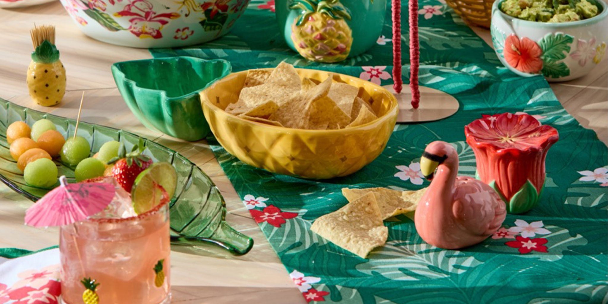 Decorative table setting with tropical-themed tablecloth, drinks, and snacks on a wooden surface.